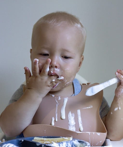 blonde haired baby boy wearing a baby bib eating yogurt off his fingers